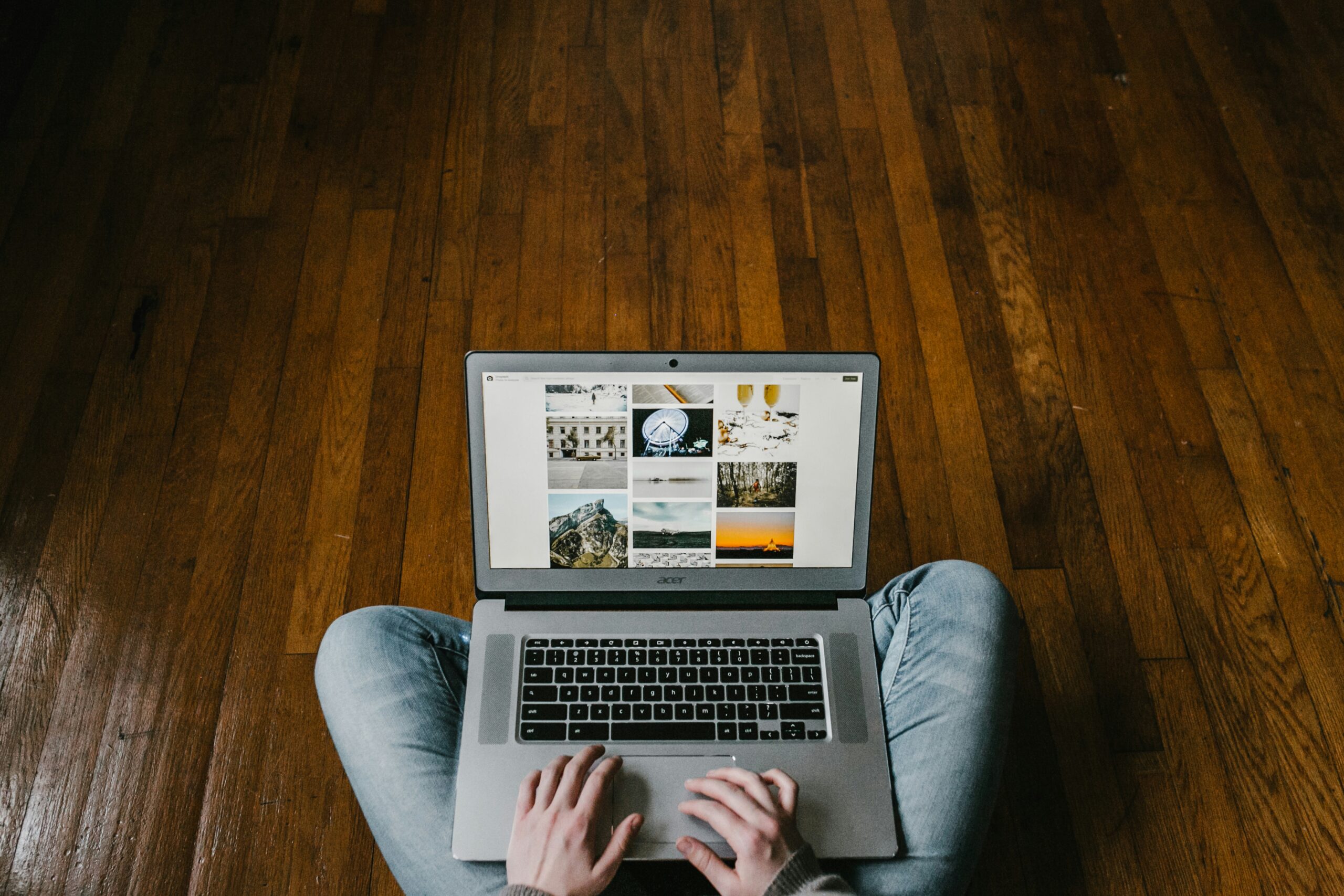 Top-down view of someone in jeans sitting cross-legged on dark hardwood flooring, typing on a laptop computer.