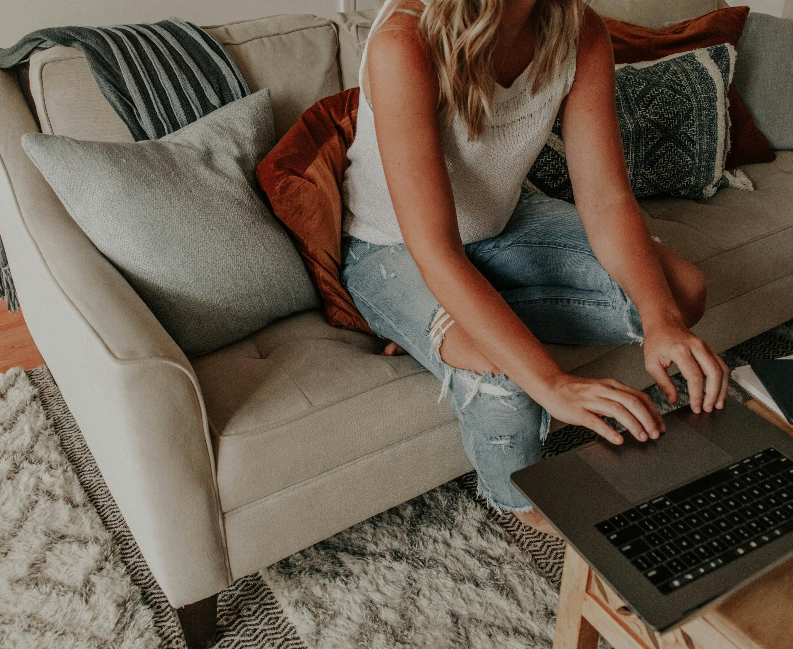 A woman sitting on a beige couch working on a laptop, representing the kind of part time side hustles that fit around a regular job